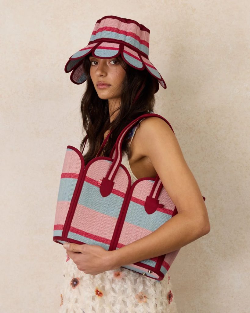 Woman in a pink and blue striped sunhat and matching striped tote bag posing against a beige wall.