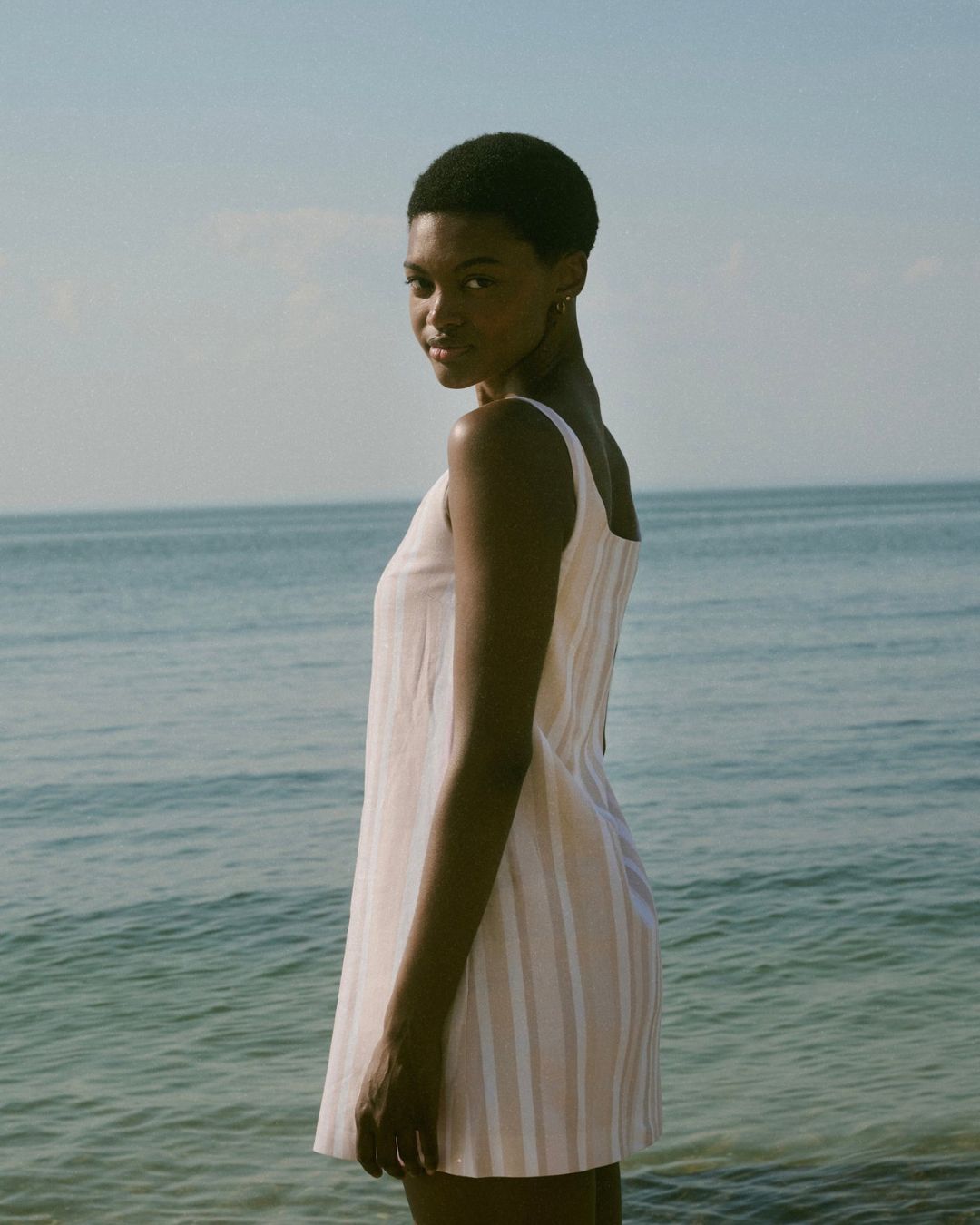Coco Shop Cappie Mini Dress Beige Stripe MHN S 26 | Michael Hayes Newport | Michael Hayes Newport Woman with short hair in a light striped sundress standing at the shoreline, looking back at the camera at the beach.