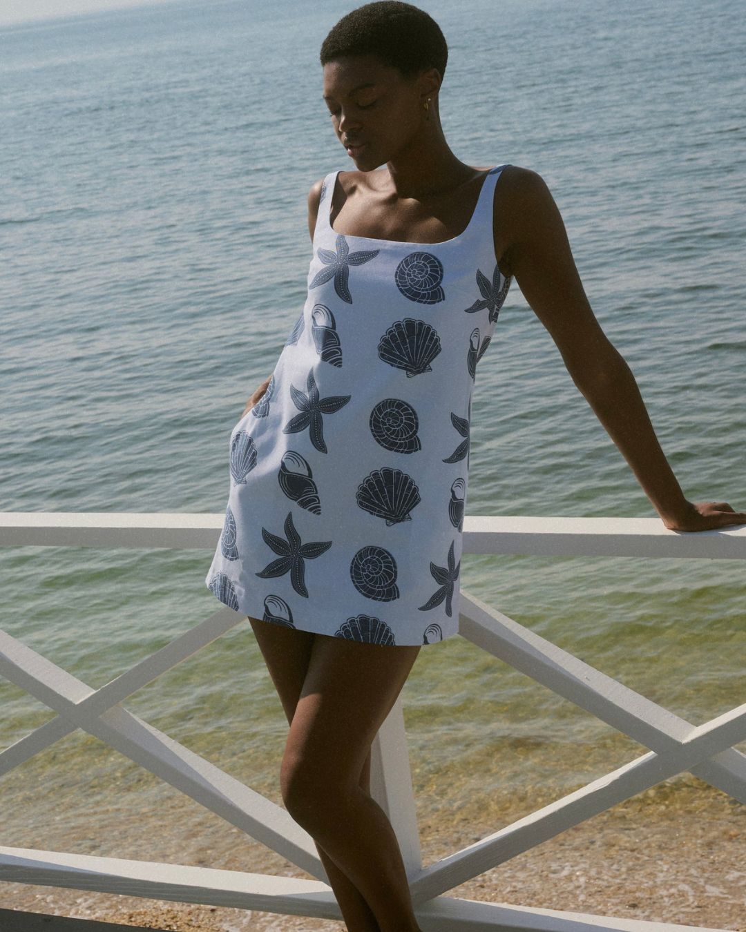 Coco Shop Cappie Mini Dress Blue Large Shell MHN S26 | Michael Hayes Newport | Michael Hayes Newport Woman in a light blue seashell-print dress leans on a white railing with the ocean in the background while standing on a beachside deck.
