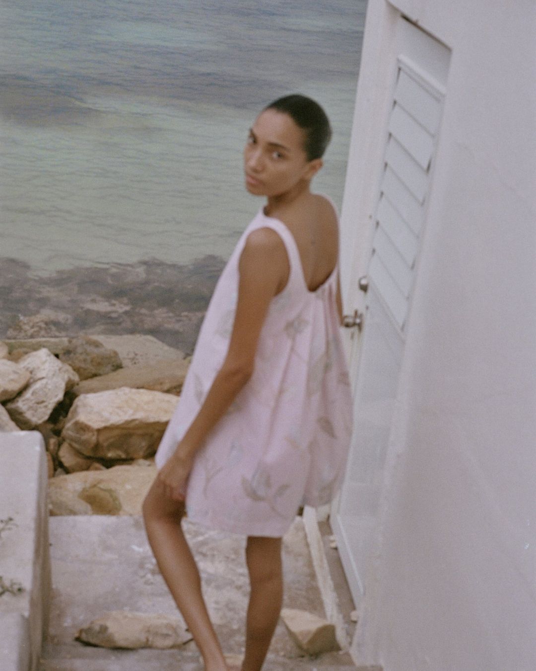Coco Shop Pleated Back Mini Dress Rose Peace Lily MHN S26 | Michael Hayes Newport | Michael Hayes Newport Woman in a pale pink sleeveless dress leans against a white wall by a rocky shore, with calm sea in the background, looking over her shoulder.