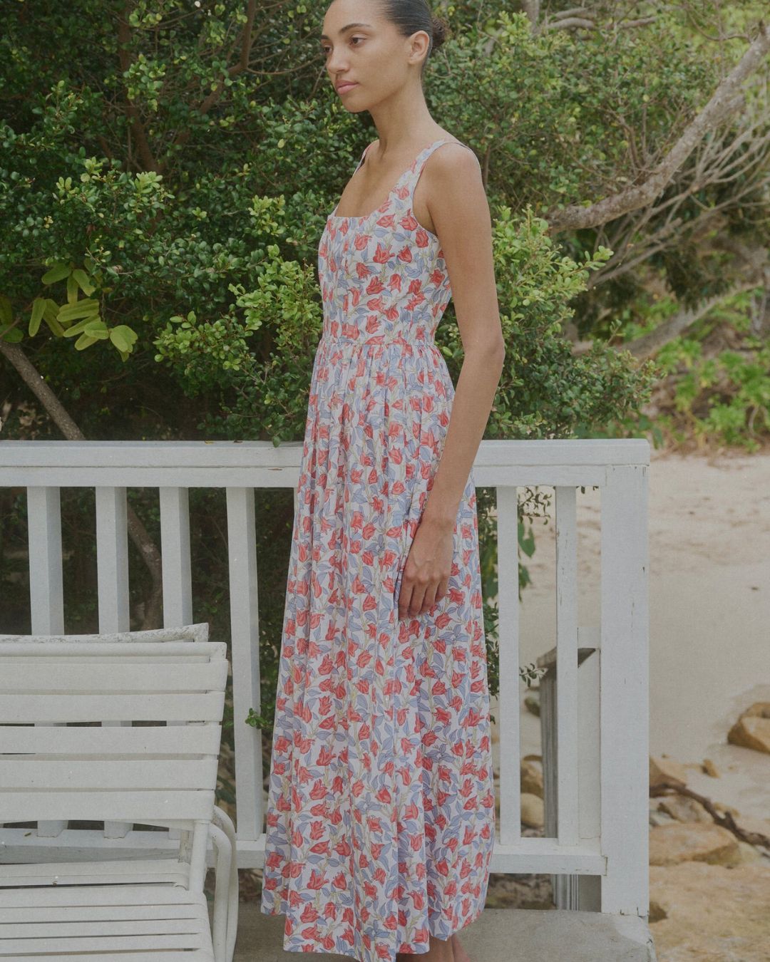 Coco Shop Scoop Neck Dress Red Bougenvillea MHN S26 | Michael Hayes Newport | Michael Hayes Newport Woman in a pastel floral maxi dress stands facing left beside a white railing near a sandy beach.