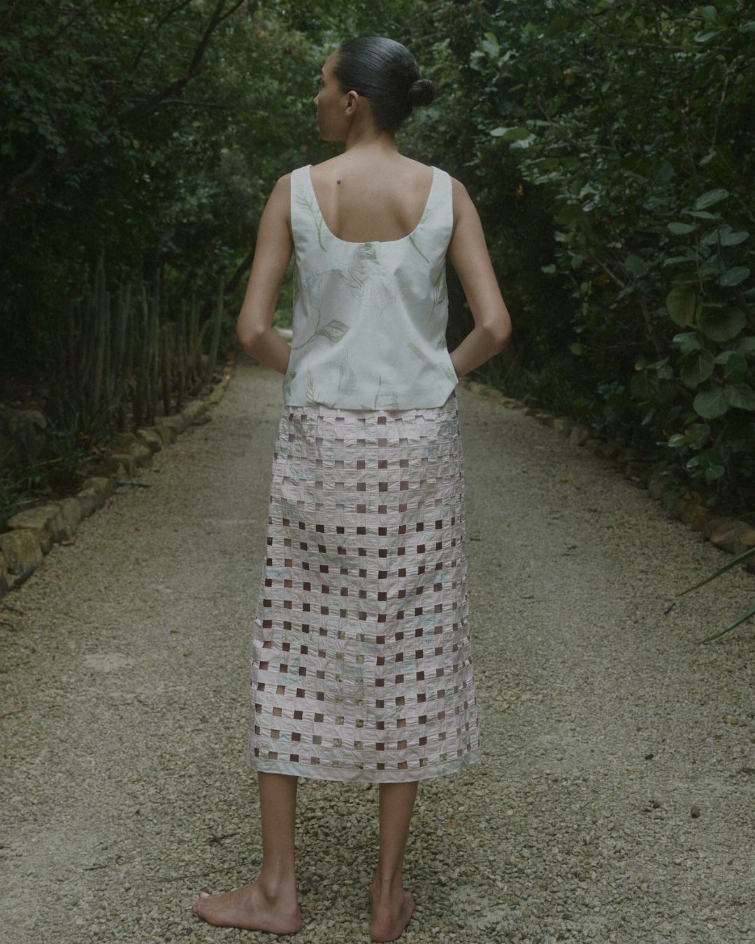 Coco Shop basket skirt at MHN | Michael Hayes Newport | Michael Hayes Newport Person standing barefoot on a gravel garden path, viewed from behind, wearing a light sleeveless top and a white lattice skirt.