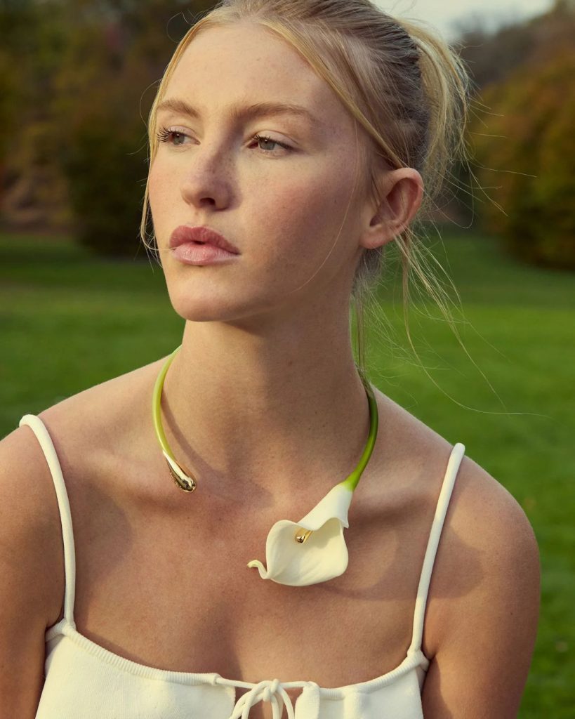 Young woman outdoors wearing a white sleeveless top and a green necklace with a white calla lily pendant.