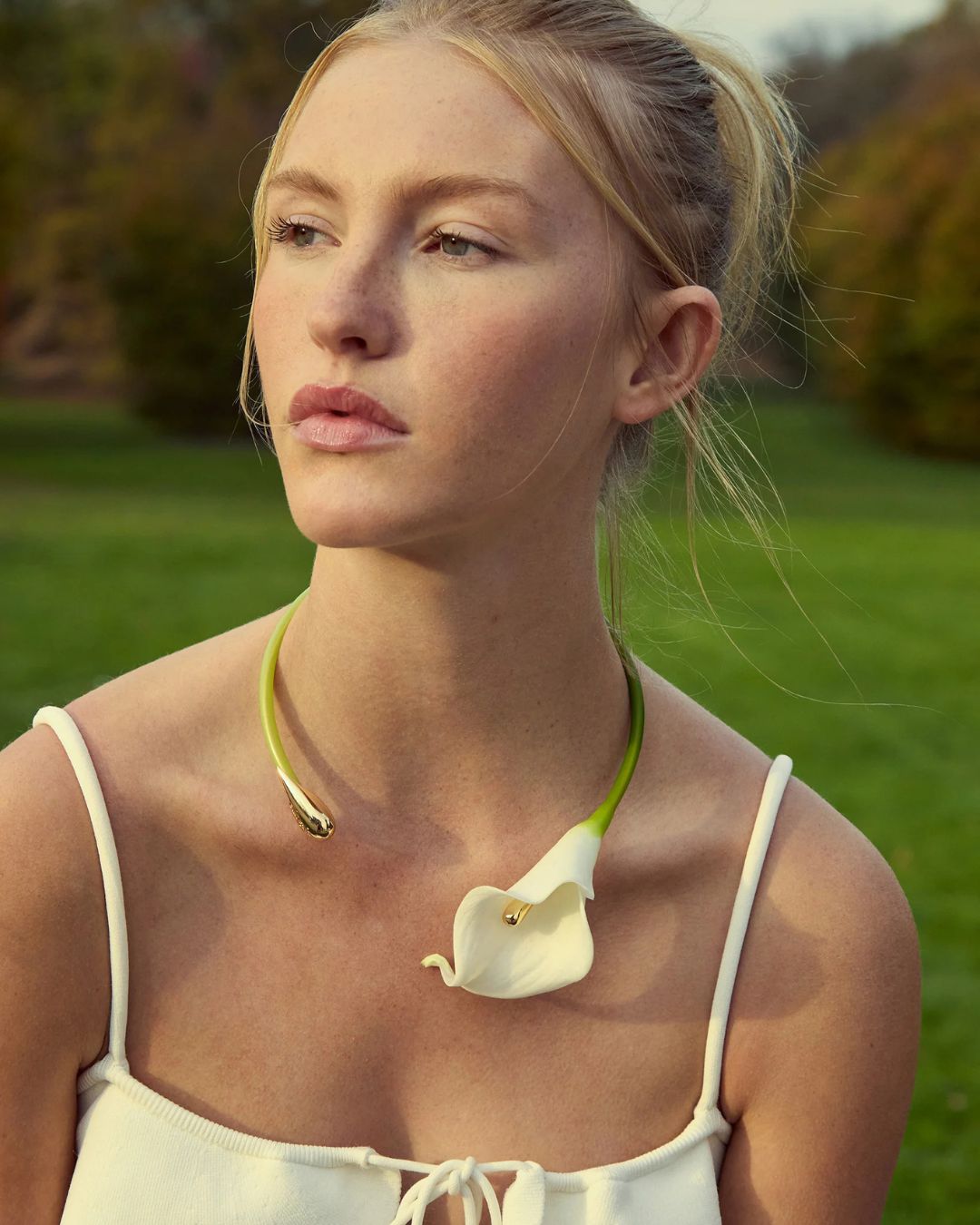 Young woman outdoors wearing a white sleeveless top and a green necklace with a white calla lily pendant.