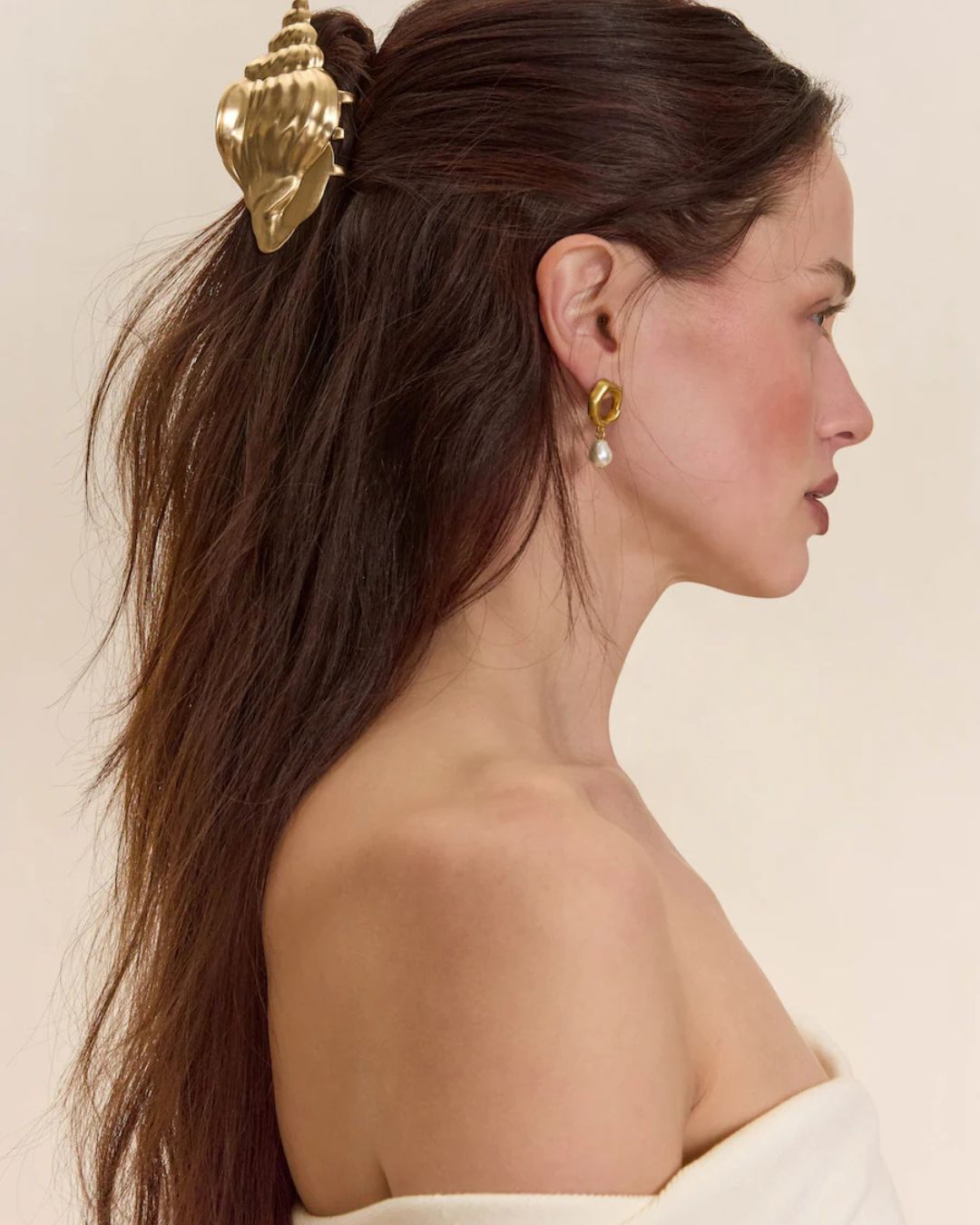 Cult Gaia Seashell Hair Clip | Michael Hayes Newport | Michael Hayes Newport Side-profile of a woman with long brown hair secured by a large gold seashell clip, wearing a gold hoop earring with a pearl drop.