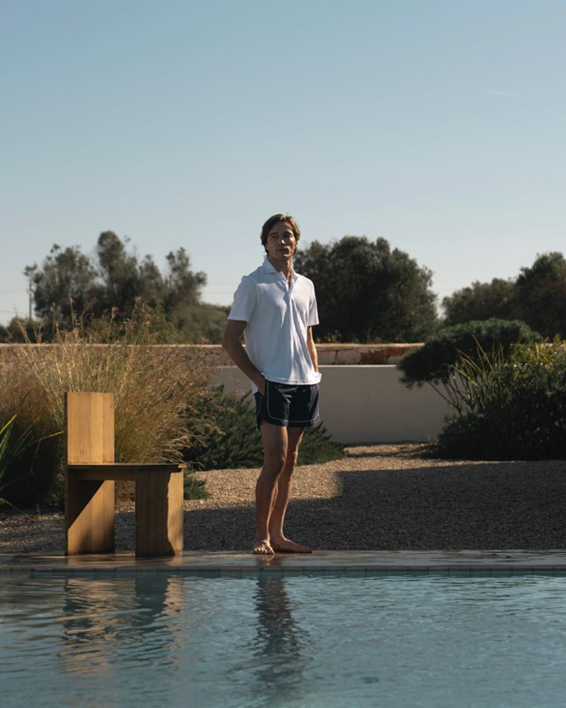Man standing barefoot at the edge of a swimming pool in a sunny backyard, wearing a white polo and dark shorts.