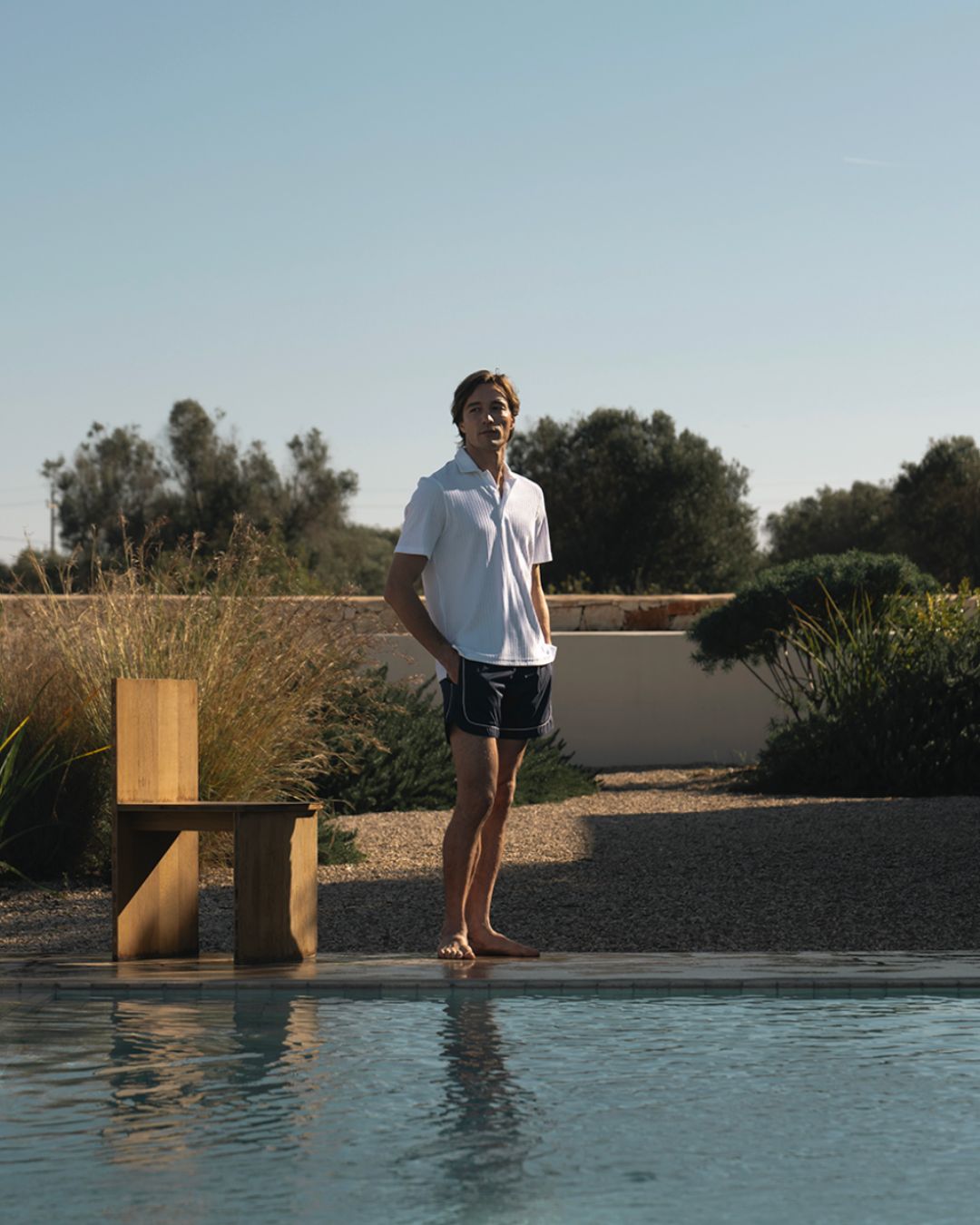 Man standing barefoot at the edge of a swimming pool in a sunny backyard, wearing a white polo and dark shorts.
