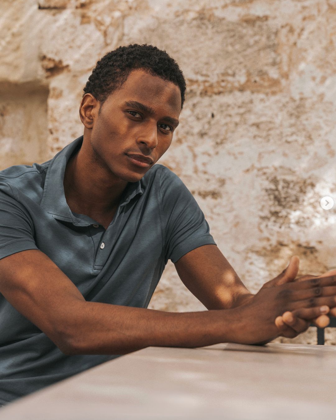Young man seated at a table outdoors, arms resting on the surface, looking toward the camera with a neutral expression against a weathered wall backdrop.