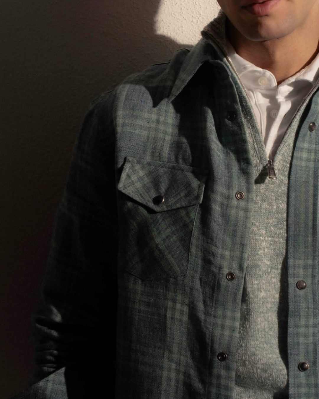 Close-up of a person wearing a green plaid button-up shirt over a white polo, showing the chest pocket and zipper detail, against a textured wall.