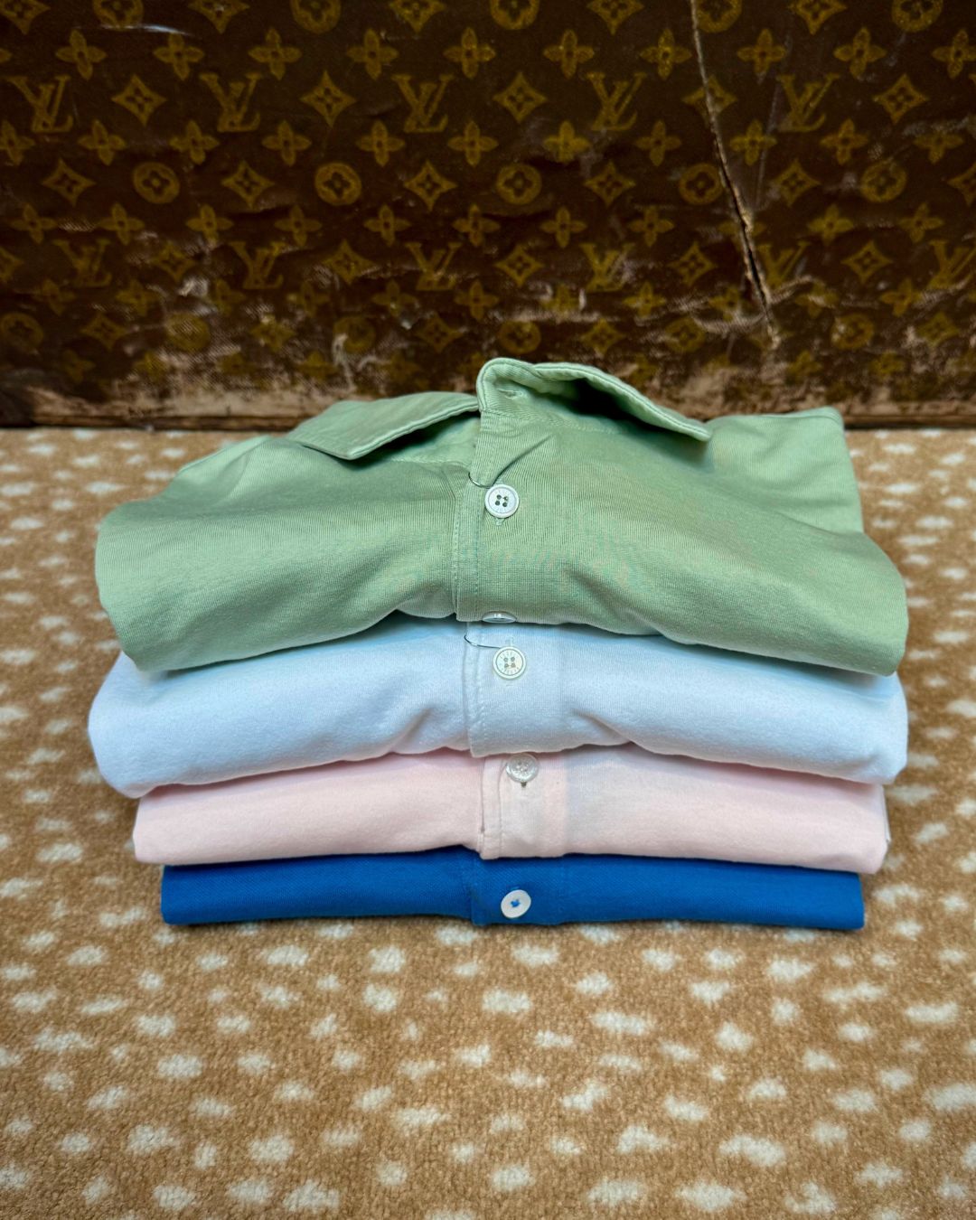 Stack of four folded pastel dress shirts in green, light blue, pink, and royal blue on a patterned carpet backdrop.