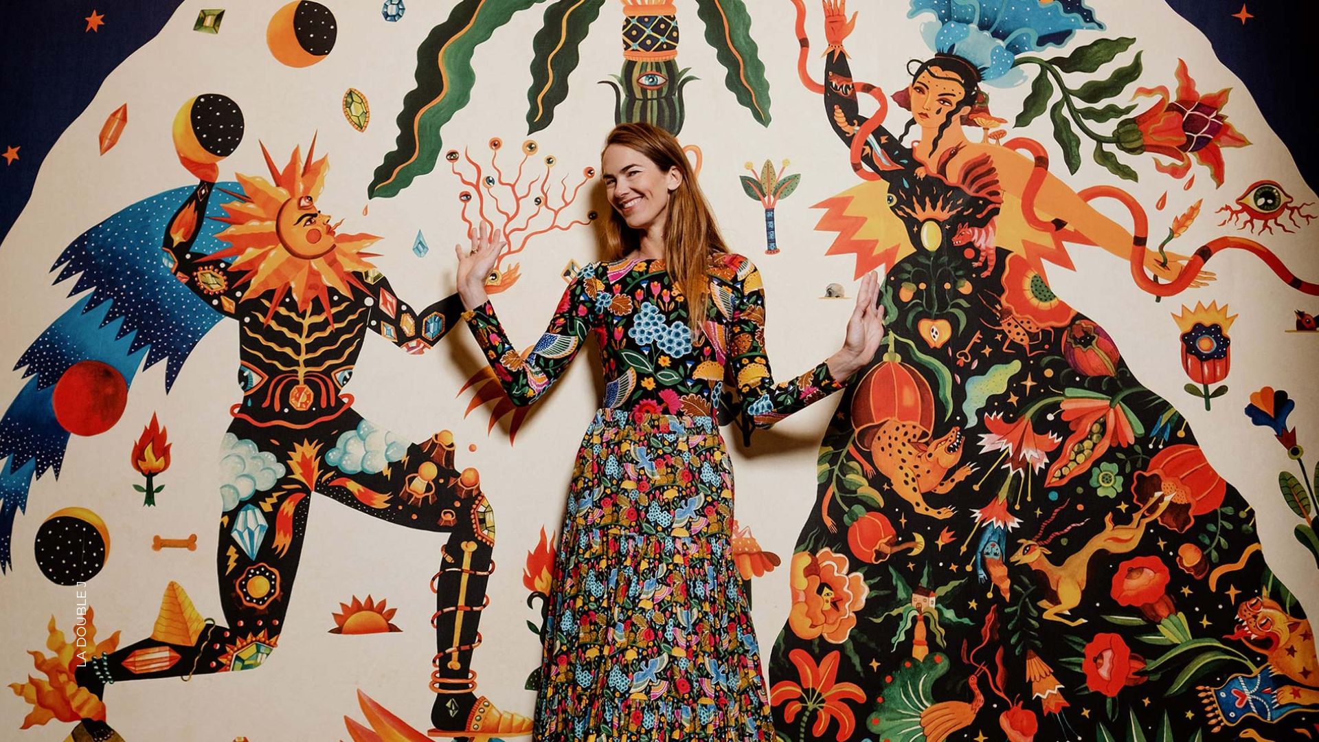 Woman in a vibrant floral dress posing in front of a large fantasy mural with colorful figures and abstract shapes